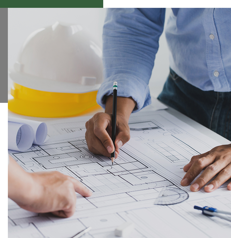 Two people work on architectural blueprints at a table. One holds a pencil over the plans, and the other points. A white hard hat and rolled-up plans are in the background.